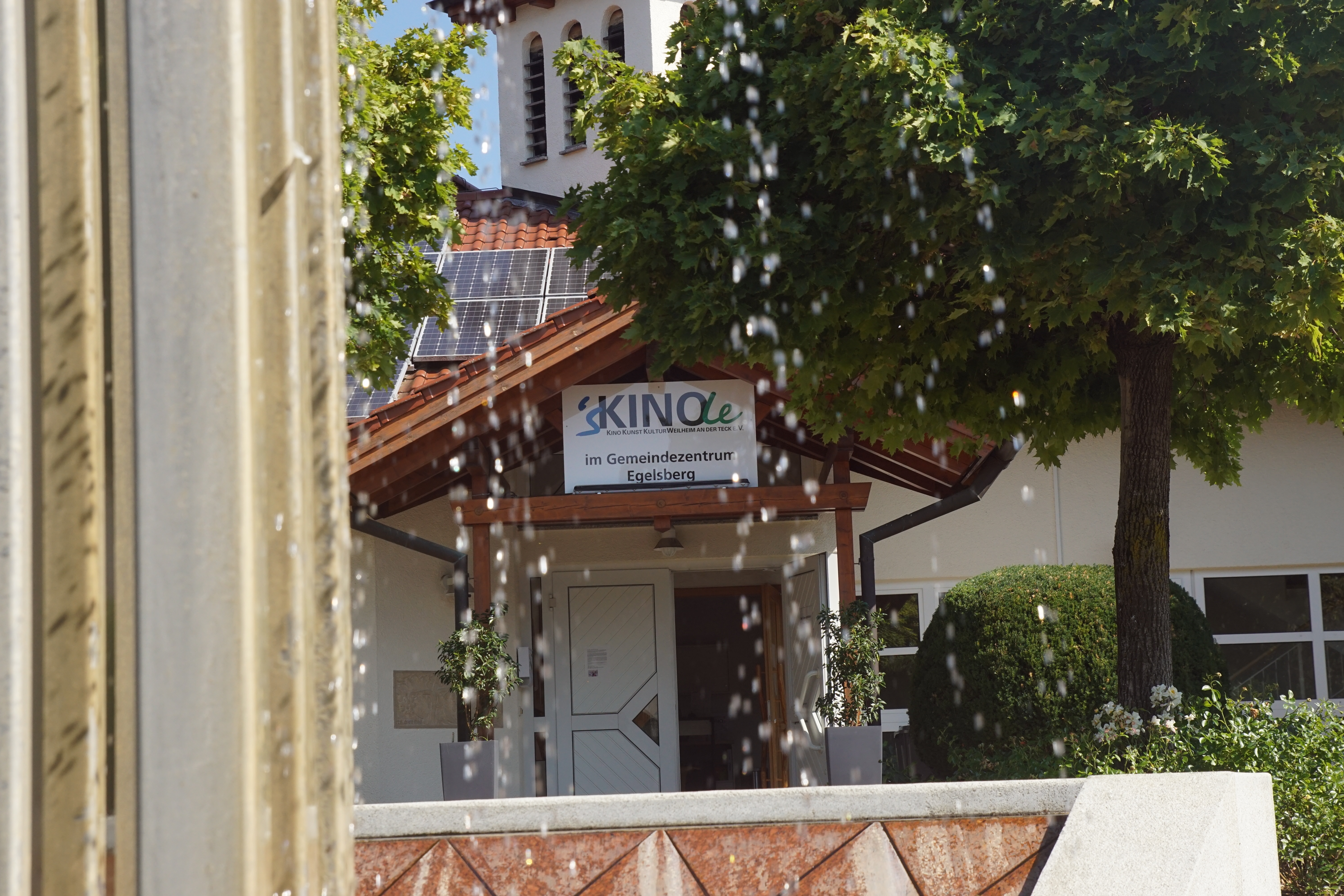 Foto des Eingangs vom Kinole - im Vordergrund der angeschnittene Brunnen, dahinter das spitze Eingangsdach mit roten Ziegeln. Unter dem Dach hängt ein weißes Schild mit der Aufschrift "sKinole". Kinole in Weilheim am Gemeindezentrum Egelsberg