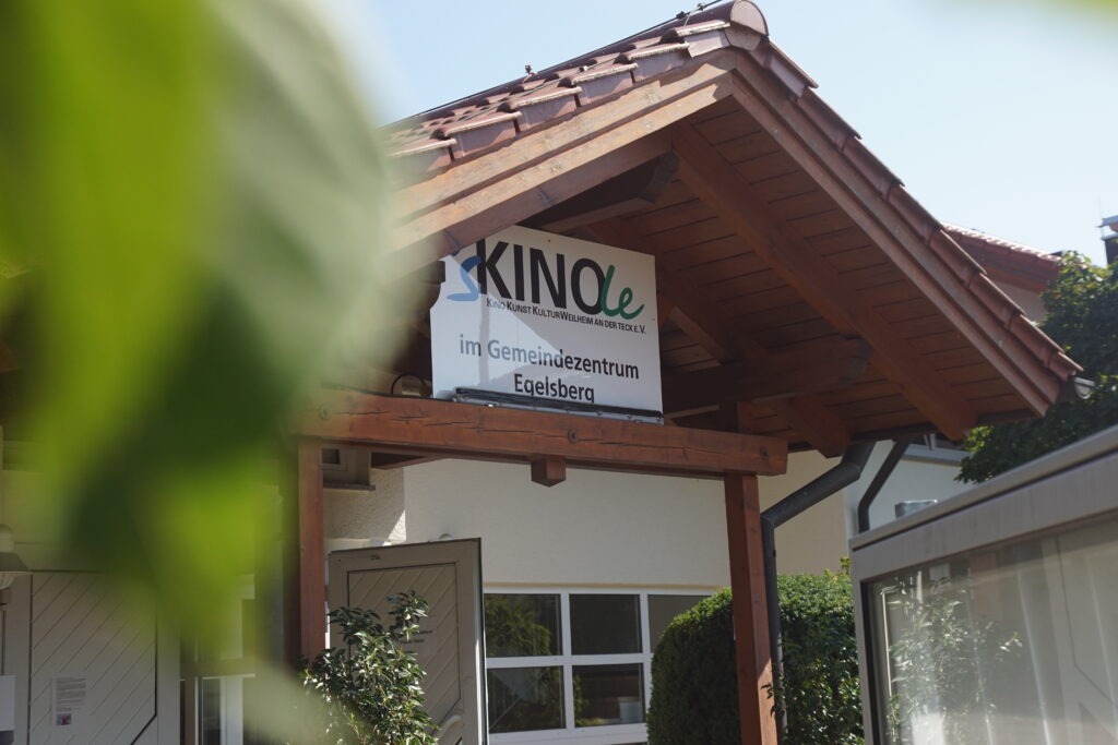 Gebäude-Eingang, spitzes mit Ziegeln bedecktes Dach über dem Eingang. Auf einem Schild über dem Eingang ist der Schriftzug vom Kinole. Besuchen Sie jetzt unser gemütliches Kinole in Weilheim an der Teck.